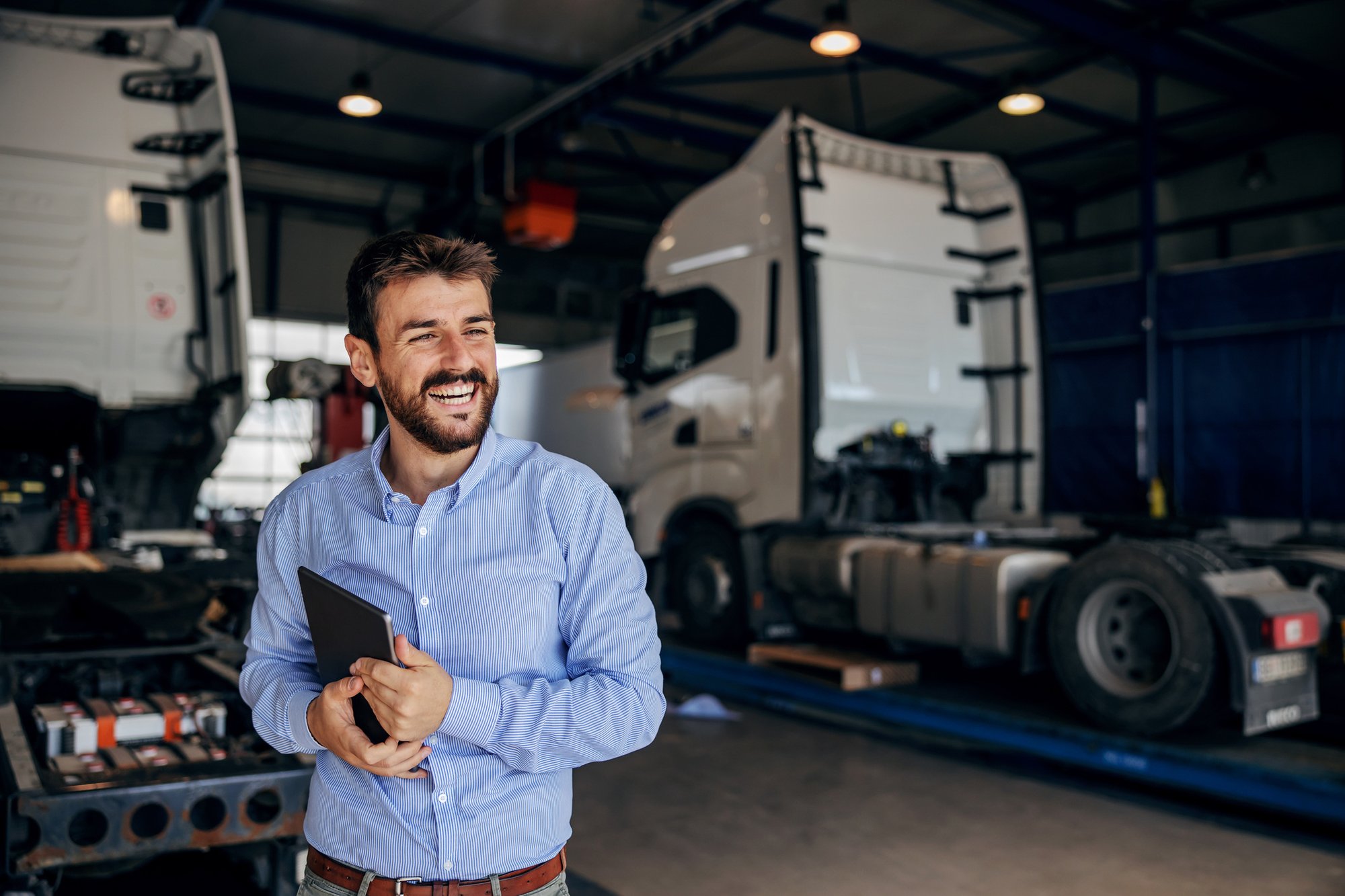 Smiling-chief-standing-in-auto-park-and-holding-tablet.-In-background-are-trucks.-Firm-for-import-and-export.-1285071058_2125x1416