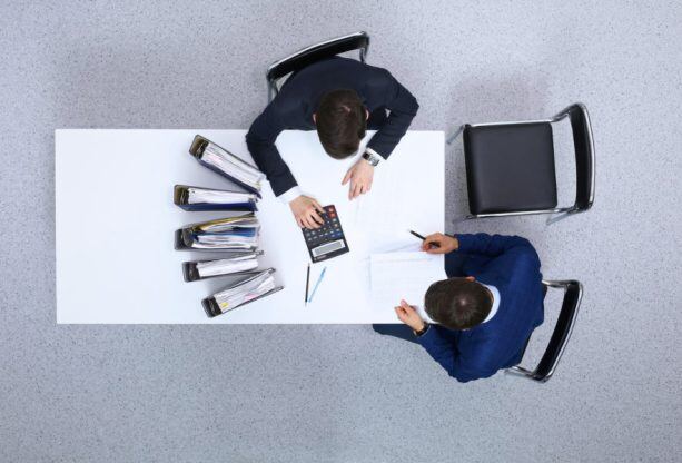 Two-businessmen-sitting-at-the-table-view-from-above.-Bookkeeper-or-financial-inspector-making-report-calculating-or-checking-balanc-952705860_1258x838-613x416